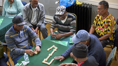 A group of men are sat around a table playing dominoes at a Coffee Morning. Some people are watching the game.