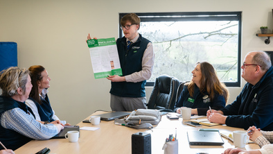 Group of people sat around a table having a meeting. One person is standing holding an A3 poster.