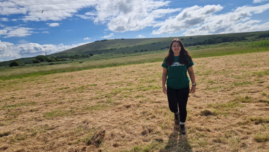 A person in a green Macmillan T-shirt is walking across a grassy field. In the background are rolling green hills and a slightly clouded sky.