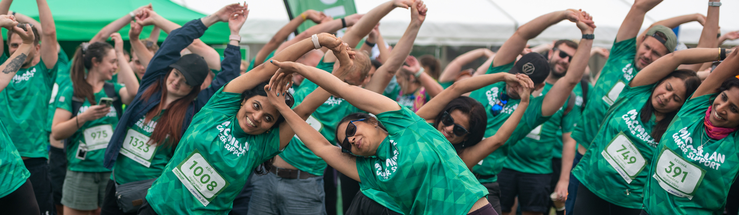 A large group of people are all bent over to the side stretching. They are wearing bright green Macmillan branded shirts and standing outside on a grassy field.