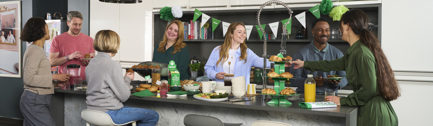 A group of people around a kitchen island for a Coffee Morning at home. The island has different delicious treats and drinks scattered across it.