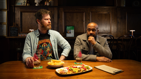 Two men are sitting side by side at a table in a pub. The man on the left is looking over to the man on the right. On the table are drinks and food. 