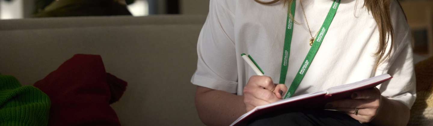 A person is sat down on a sofa and is writing in a notebook. They have long brown hair and are wearing a white T-shirt and a Macmillan lanyard. 