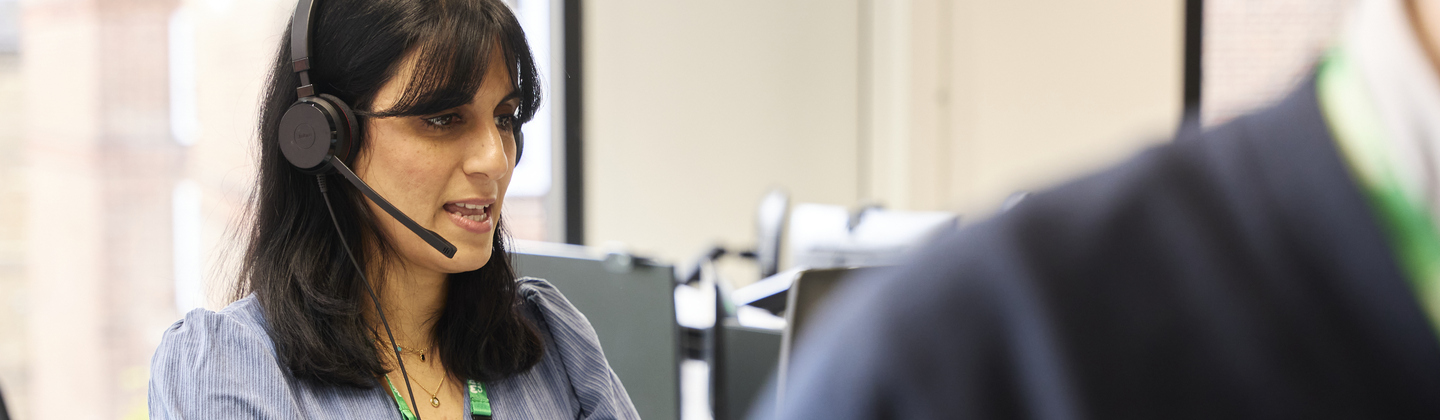 Sunni is standing at a computer desk. She is wearing a headset and speaking. She has dark shoulder length hair and is wearing a blue and white stripped blouse.