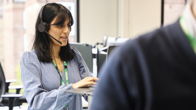 Sunni is standing at a computer desk. She is wearing a headset and speaking. She has dark shoulder length hair and is wearing a blue and white stripped blouse.