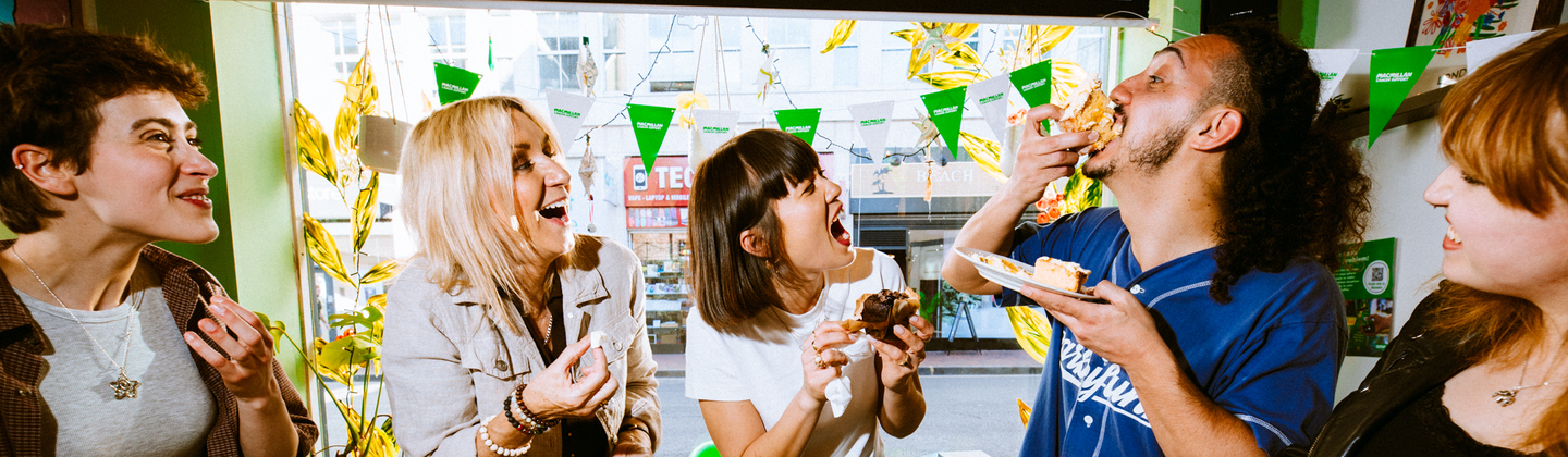 A group of people are standing around a table full of delicious nibbles at a Coffee Morning. One person is taking a bite of a massive slice of a moreish cake whilst the others are watching on.