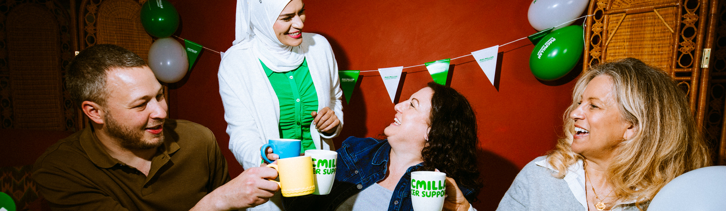A group of four people are gathered around a table for a Coffee Morning. They are each holding a mug and are toasting. The table has different yummy treats on it like cupcakes, muffins and a bowl of fruit.