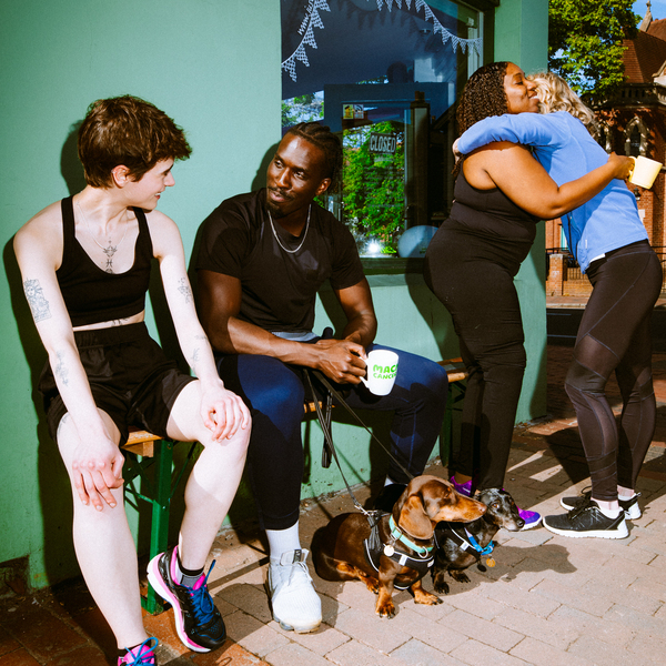 People are gathered outside a café enjoying a sunny day and a Coffee Morning. They are wearing active wear. Two people are hugging, whilst the other two are in mid conversation. 