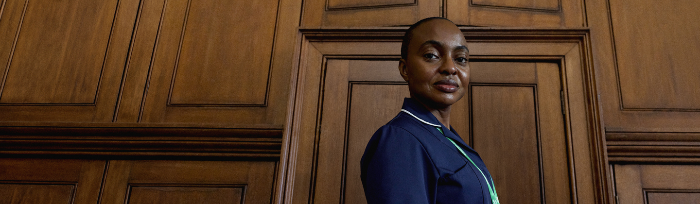 A Macmillan nurse standing from the side and looking towards the camera. They are standing in a room with wood panelling. They are wearing a dark blue NHS uniform.