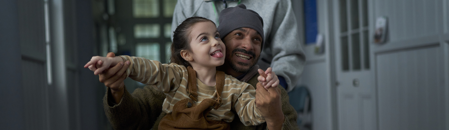 A father is sitting down in a wheelchair that is being pushing through a corridor. His daughter is in his lap and they are holding hands in the air. 