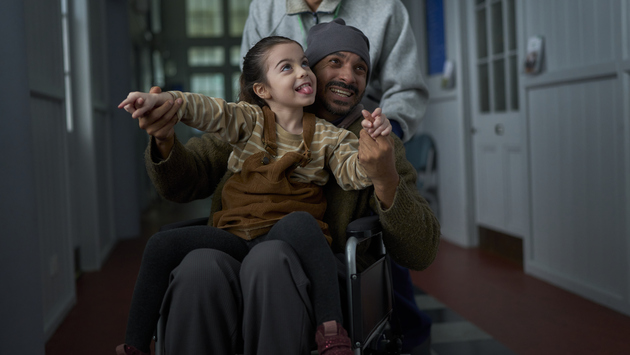 A father is sitting down in a wheelchair that is being pushing through a corridor. His daughter is in his lap and they are holding hands in the air. 