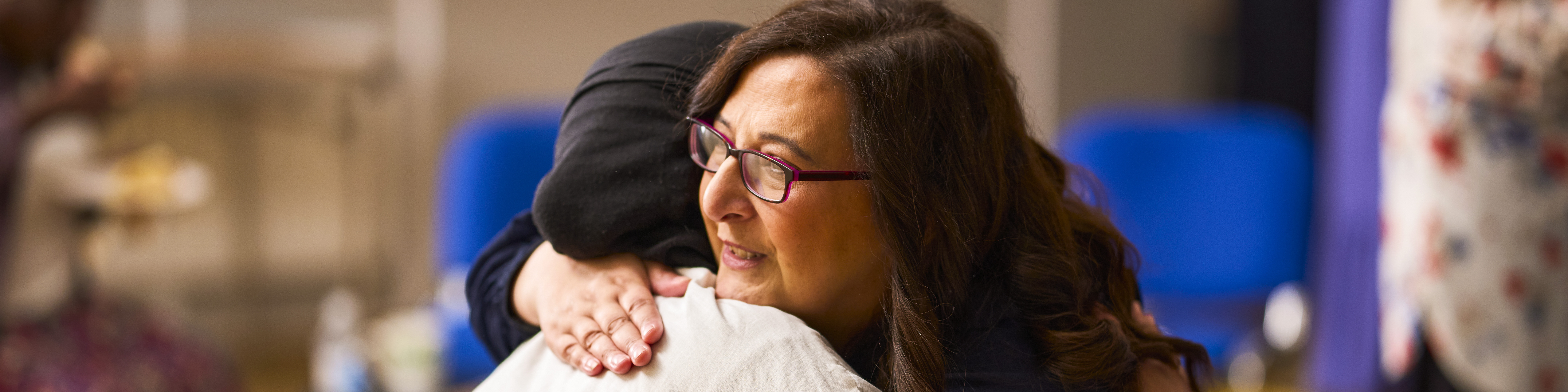 Two women hug. One has dark hair and is wearing glasses the other is wearing a headscarf and light colour shirt.