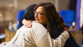 Two women hug. One has dark hair and is wearing glasses the other is wearing a headscarf and light colour shirt.
