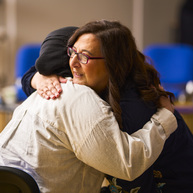 Two women hug. One has dark hair and is wearing glasses the other is wearing a headscarf and light colour shirt.