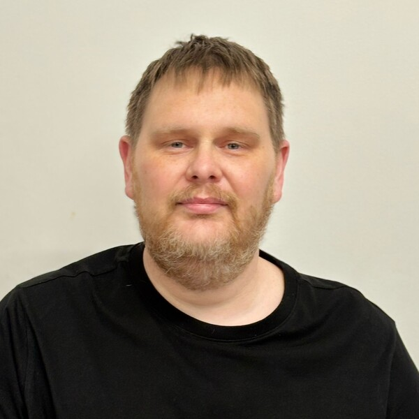 A white man with light brown hair and a beard with a black t shirt on.