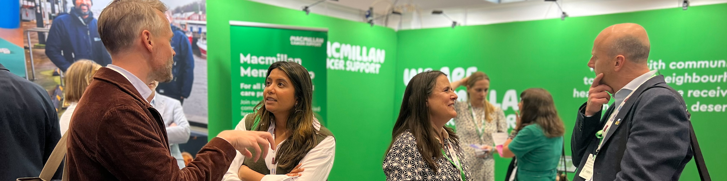 People speaking at a Macmillan Cancer Support stand at an indoor event, with information materials displayed on a green counter.