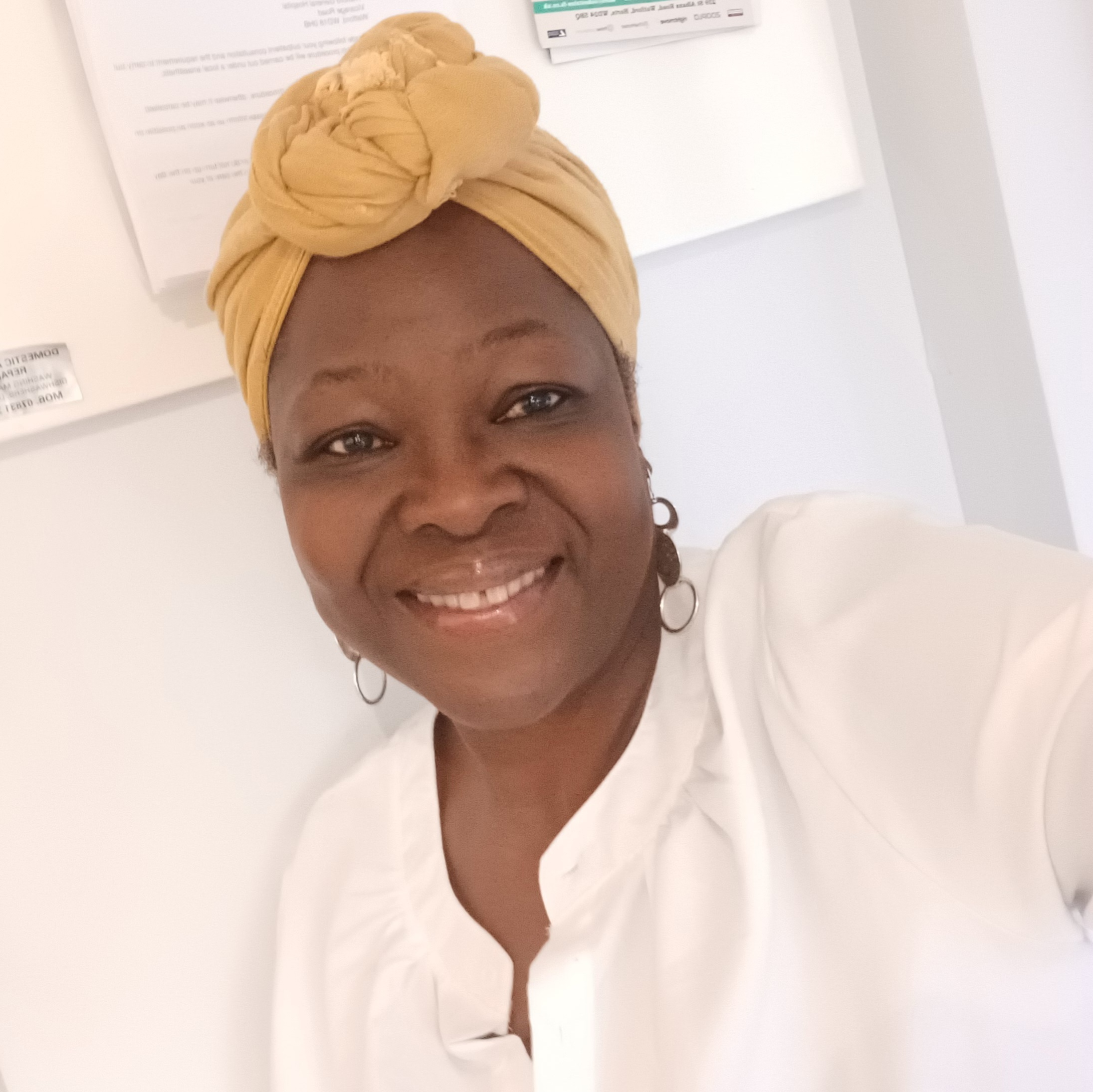 Woman smiles at camera, she is wearing a white shirt and mustard coloured headwrap