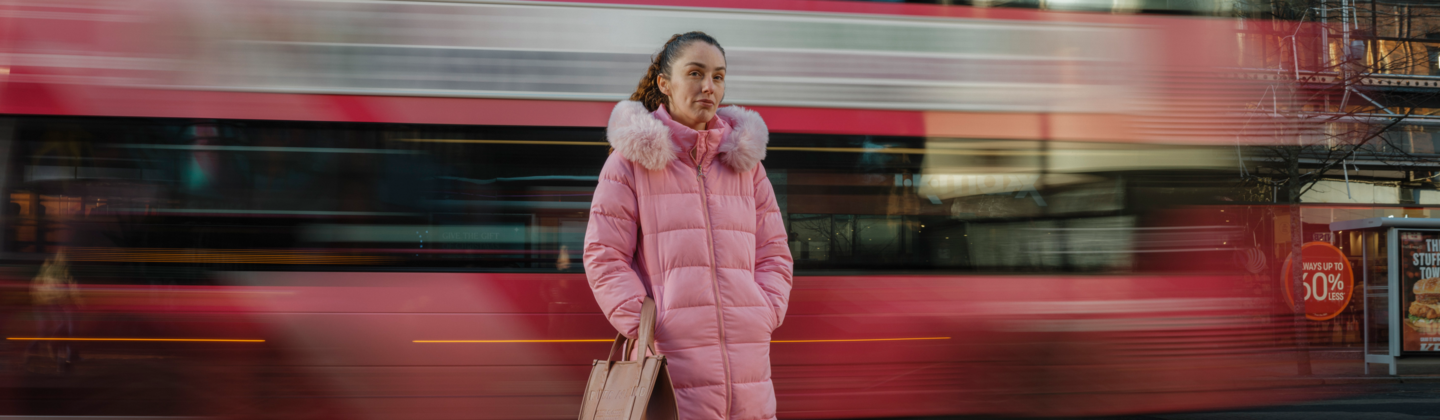 A person in a long pink coat stands at a bus stop while a bus passes by on a city street.