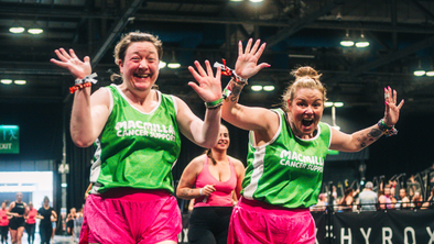 Two women wearing pink shorts and green Macmillan running vests waving at the camera during a HYROX event
