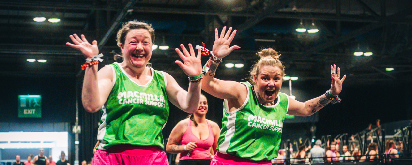 Two women wearing pink shorts and green Macmillan running vests waving at the camera during a HYROX event