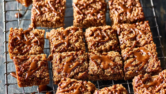 A batch of rich, golden-brown flapjacks cut into neat squares and arranged on a cooling rack. Each piece has a slightly crisp edge and a chewy oat texture, with a glossy drizzle of mocha icing zigzagged across the top. The icing, made with Camp Coffee, adds a deep coffee aroma and a hint of sweetness. These indulgent treats look homemade and comforting — perfect for coffee lovers seeking a twist on a classic bake.