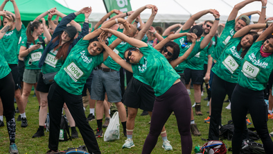 A large group of people are all bent over to the side stretching. They are wearing bright green Macmillan branded shirts and standing outside on a grassy field.