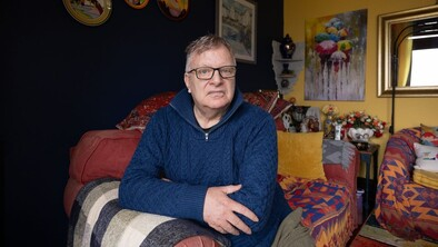 A white man with glasses and a blue jumper sat on his sofa.