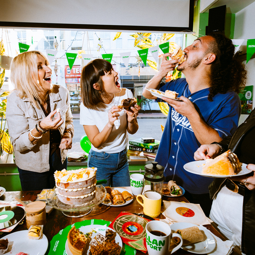 A group of people are standing around a table full of delicious nibbles at a Coffee Morning. One person is taking a bite of a massive slice of a moreish cake whilst the others are watching on.