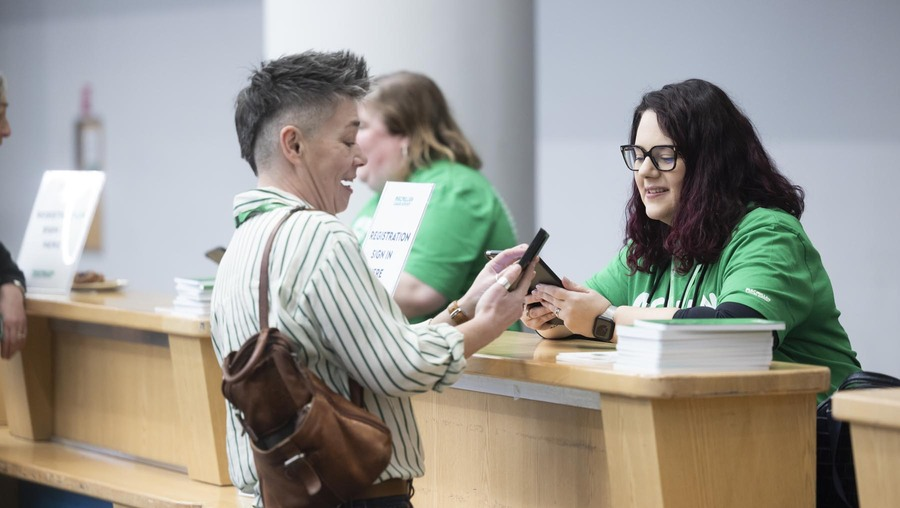 A person is standing behind a desk. They are speaking to someone who is checking their mobile. They appear to be at an event.