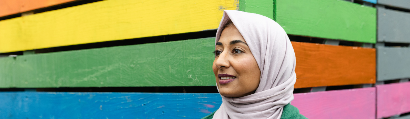 Saliha Ahmed from the Macmillan Bristol Cancer Champions project. Saliha is standing outside in front of a building painted in rainbow colours. She is wearing a grey headscarf, grey top and a Macmillan branded coat.