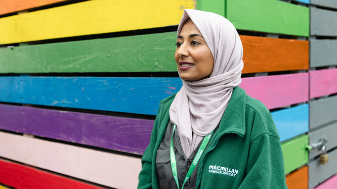 Saliha Ahmed from the Macmillan Bristol Cancer Champions project. Saliha is standing outside in front of a building painted in rainbow colours. She is wearing a grey headscarf, grey top and a Macmillan branded coat.