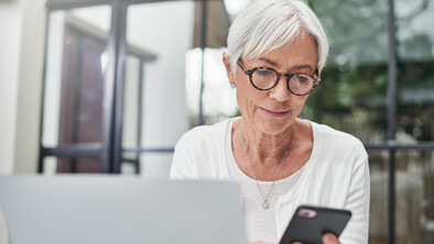 A woman with glasses and grey hair is looking at her phone. She has a laptop in front of her.