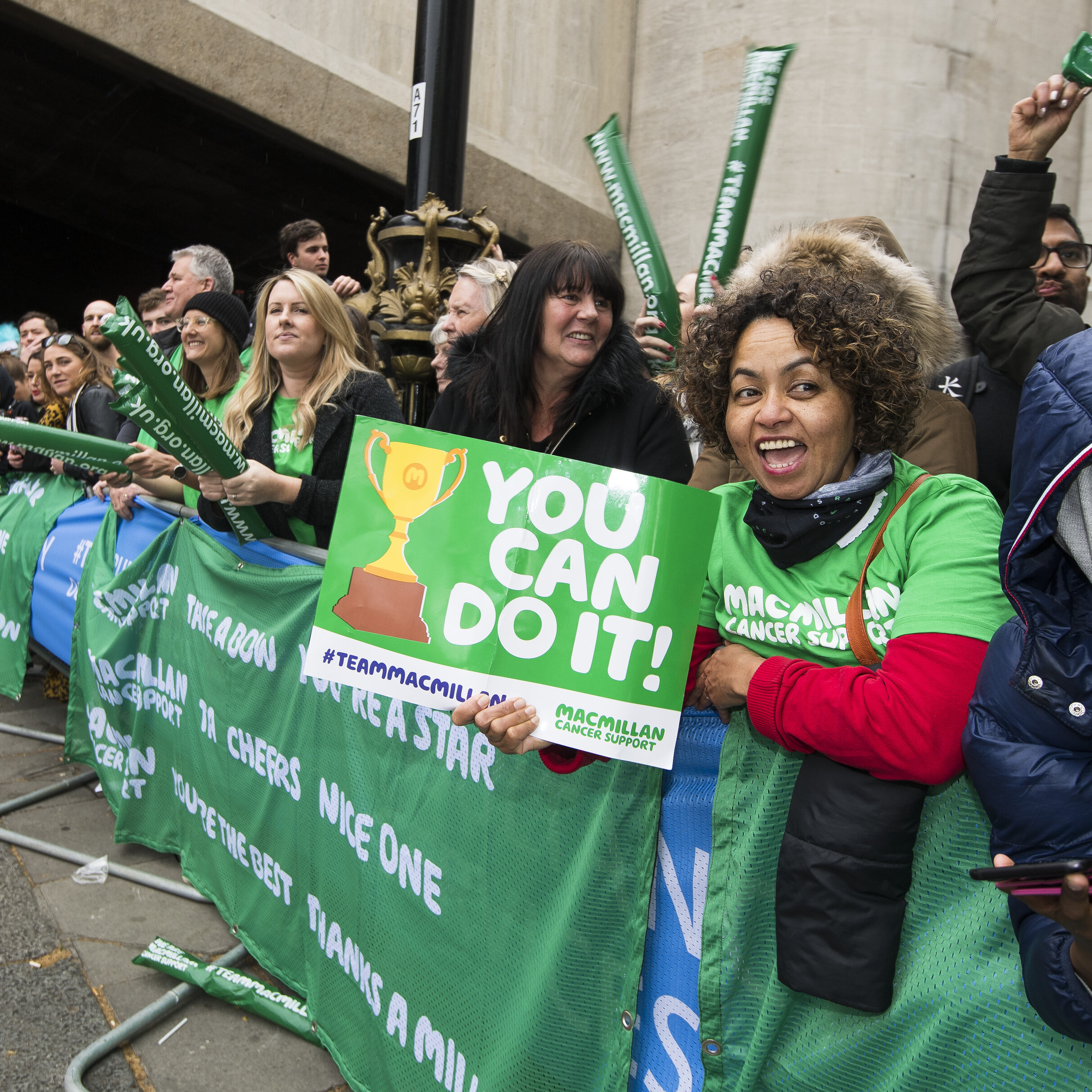 A group of volunteers are standing behind a barrier at a fundraising event. They are cheering on participants. One volunteer is holding a sign that says you can do it.