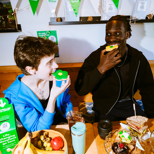 Two people are enjoying flower shaped biscuits at a Coffee Morning. In front of them is a spread including a bowl of fruit, sandwiches, sweet treats and more.