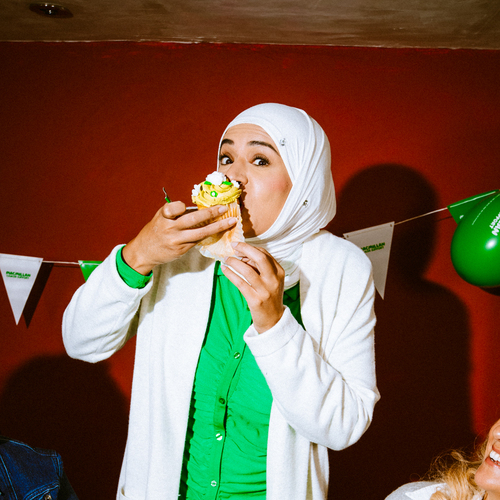 A person is enjoying a large frosted cupcake at a Coffee Morning. They are standing in between two sat onlookers. 