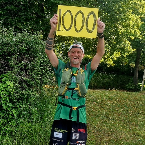 Ben is standing with his arms in the air holding a yellow sign that says 1000 on it. He is outside and is wearing different running gear. 