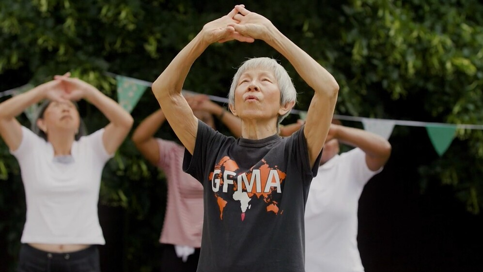 A screenshot from a video about Global Fusion Music and Arts' Coffee Morning. It includes a group of people exercising. The person in front has both arms overhead and they are stretching them.
