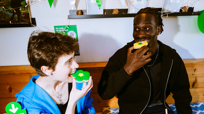 Two people are enjoying flower shaped biscuits at a Coffee Morning. In front of them is a spread including a bowl of fruit, sandwiches, sweet treats and more.