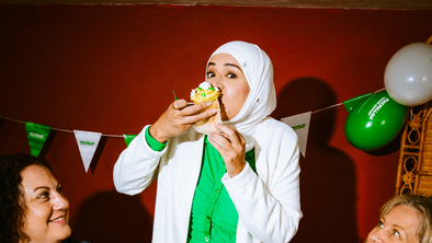 A person is enjoying a large frosted cupcake at a Coffee Morning. They are standing in between two sat onlookers. 