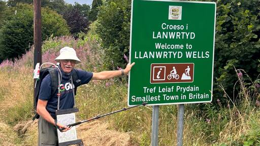 John is standing outside and learning on a green sign with information in Welsh and English stating Welcome to Llanwrtyd Wells. John is wearing a white cap, dark blue T-shirt and trousers.