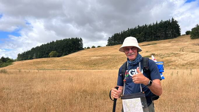 John is standing in a grassy field and giving a thumbs-up. He is wearing a white cap, dark blue T-shirt, dark hiking trousers and hiking gear. 