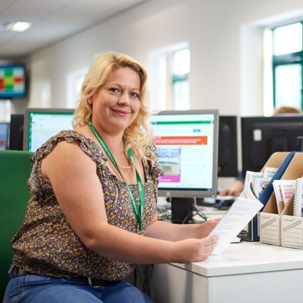 A Macmillan Support Line advisor is sitting at a desk in front of a computer and information booklets. She's smiling to the camera.
