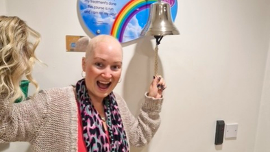 Sarah is standing next to a bell in a hospital. She is ringing it with one hand and in her other hand is a wig. Sarah is wearing a cardigan, colourful scarf and a red top.