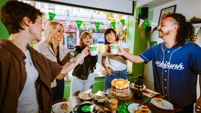A group of people gathered for a Coffee Morning. They are clinking their mugs together and doing a cheers. They are standing around a table decorated for the event.