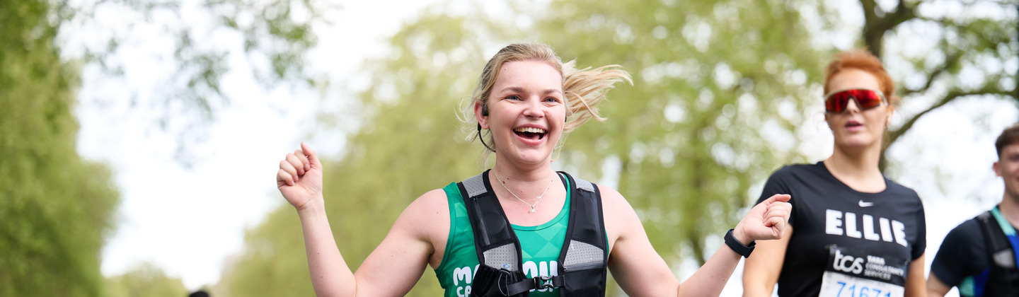 A person taking part in running event. They have their hair up in a ponytail. They are wearing a running vest, a Macmillan running top and shorts.