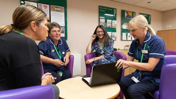 Four people sit around a small round table in a room, three in uniforms and one in casual clothing. One person uses a laptop while green Macmillan Cancer Support boards with posters and leaflets are visible in the background.