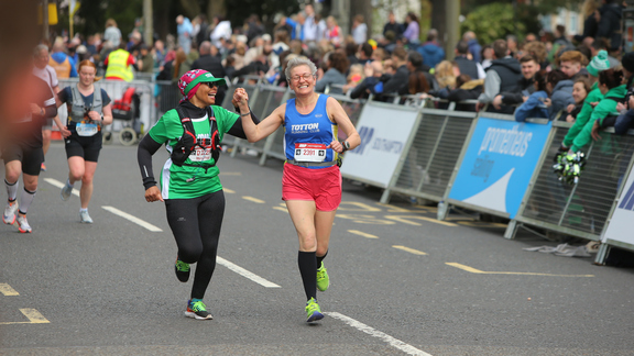 Two runners taking part in an event. They are holding hands. One runner is wearing a green Macmillan branded top. The other is wearing a blue running top.
