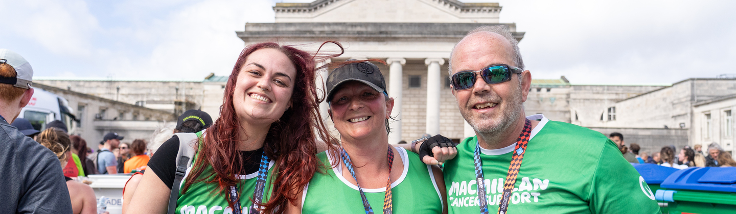 Three runners are standing next to each other after completing a running event. They are wearing green Macmillan branded running tops and are smiling.