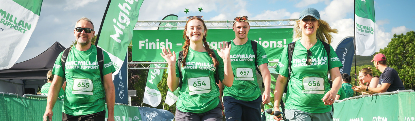 A group of four Mighty Hikers crossing the finish line. They are all wearing bright green Macmillan tops and other hiking gear.