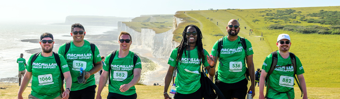 A group of Mighty Hikers trekking uphill together at the South Coast Mighty Hike. They are all wearing green Macmillan branded tops and other hiking gears. In the distance are rolling green hills and sea views of the iconic South Coast path.